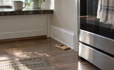 An untouched, baited wooden snap trap set against the baseboard beside a stove on a clean kitchen floor in early morning light.